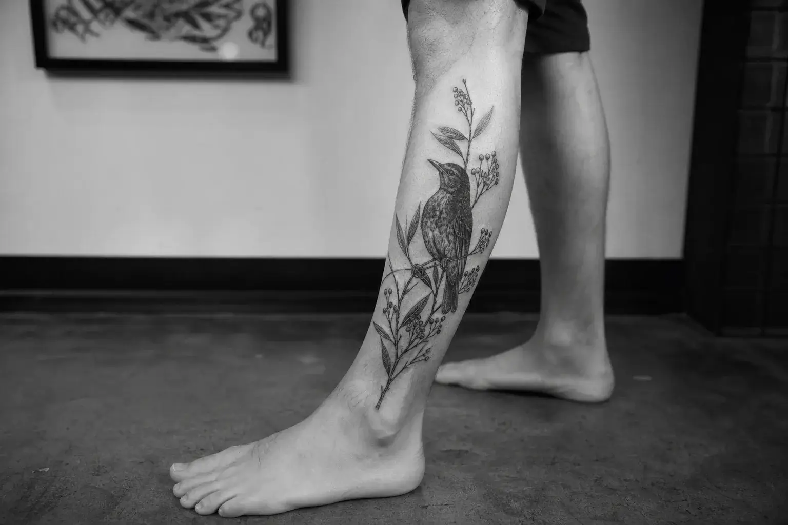 Tatouage noir et gris d’un oiseau entouré de feuillage sur un mollet